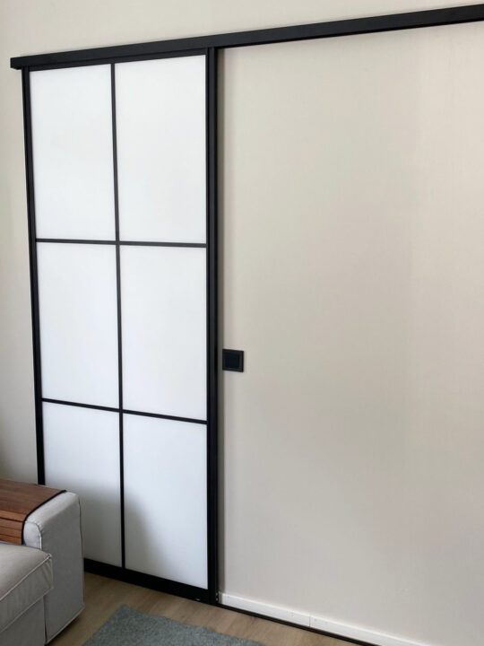 Seinä, jota vasten on yläkantoinen mustakehyksen tilanjakajaliukuovi, jonka levyt ovat valkoiset.
